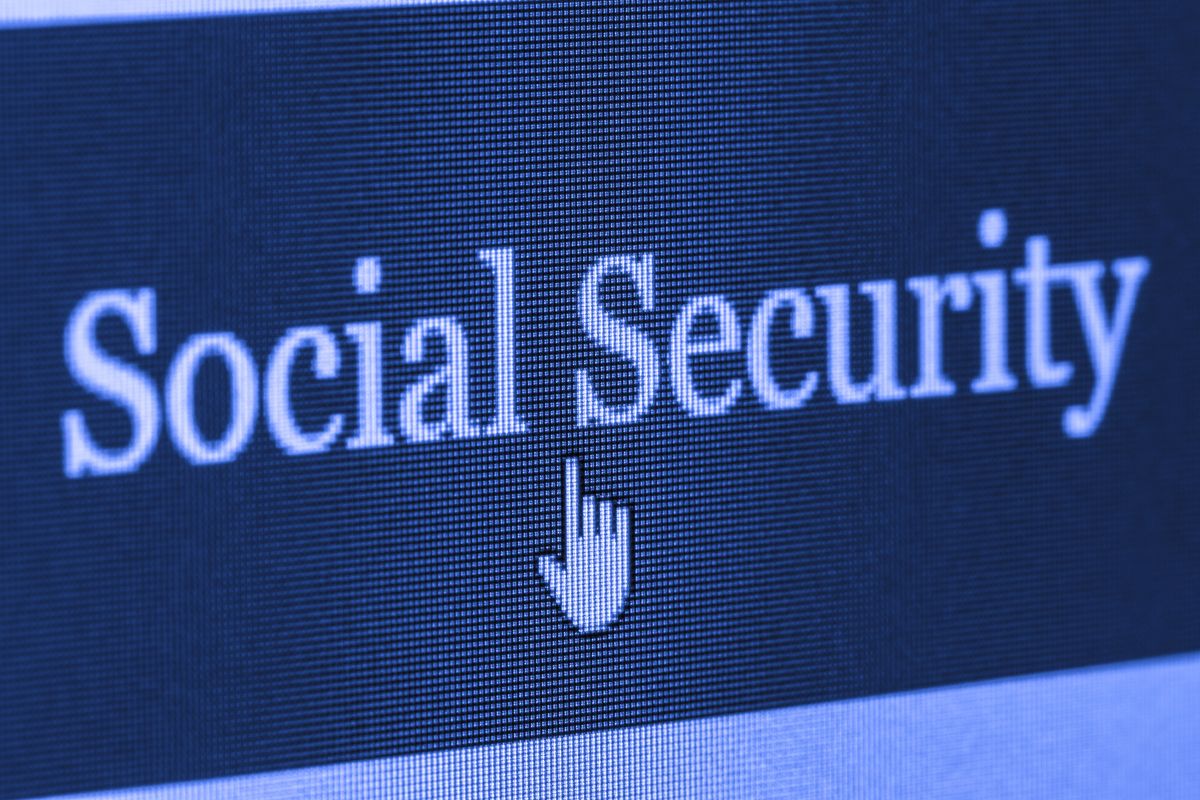 An adult sitting at a kitchen table using a laptop to sign in to a Social Security online account, with paperwork and a coffee mug nearby